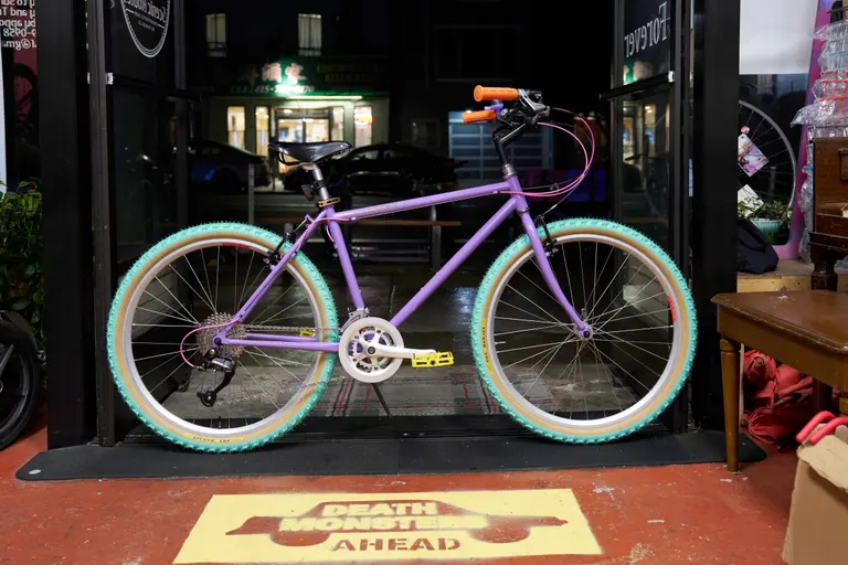 A violet bike in the bike shop with green tires, orange grips, and a dark Brooks saddle.