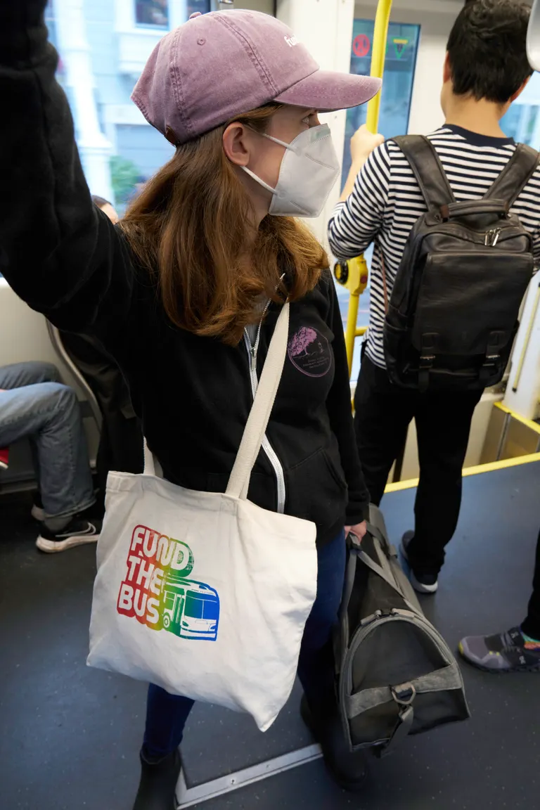 A woman, Kat, in a black sweater and plum baseball hat standing on a moving train holding a cat carrier.