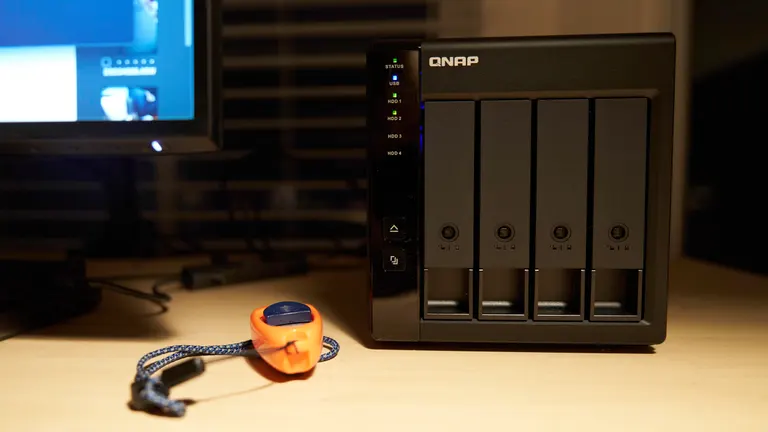 A QNAP TR-004 with four drive bays is placed on a desk. A computer monitor is partially visible in the background, and the room is dimly lit.