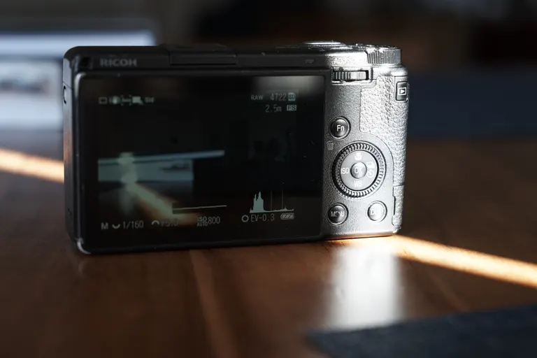 The back of a Ricoh GR III with a sliver of sunlight on the controls on its right. The LCD is on showing the dark indoor scene in front of it.