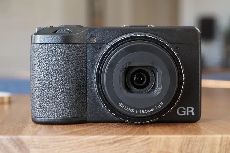 The front of a small black camera that reads "GR LENS f=18.3mm 1:2.8 GR". It is sitting on a wooden table.