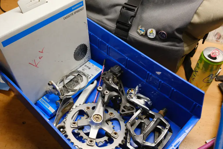 A bin containing a crankset, rear derailleur, old pedals, old front derailleur, cassette box, and chain box.