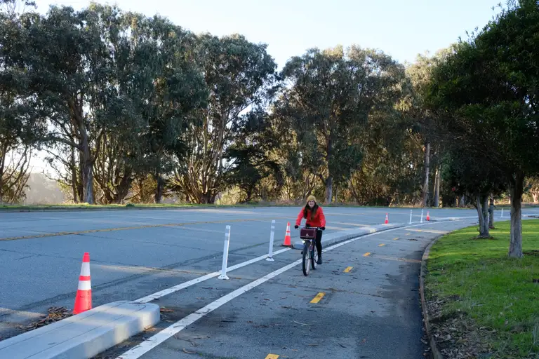 Kat riding up a two-way bike lane with concrete protection, bollars, and construction pylons. Golden hour trees and Lake Merced are behind her.