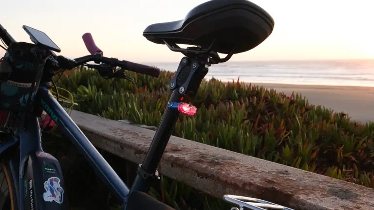 A blue e-bike against a metal railing in front of a beach sunset. A blue light is mounted on a black seatpost with a silver tensioning strap and red LED.
