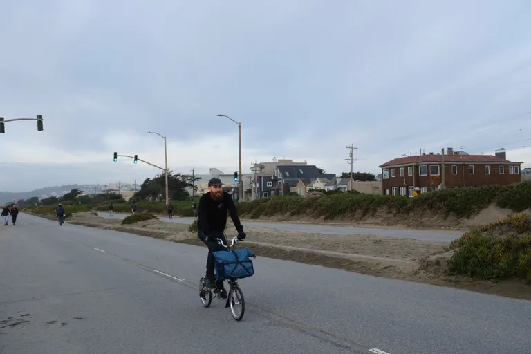 Ben riding a folding bike with a large blue bag on a cloudy day.