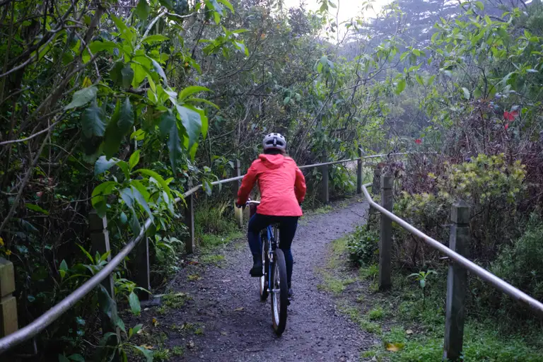 Kat biking down a gravel path with railings and foliage on each side. It's all wet and misty.
