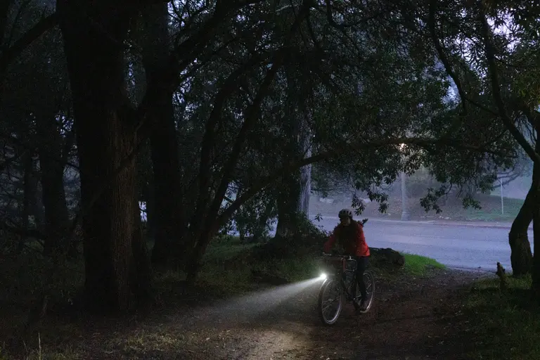 Kat biking up a dirt trail just off the road with a cone of light from her dynamo and tree leaves framing her.