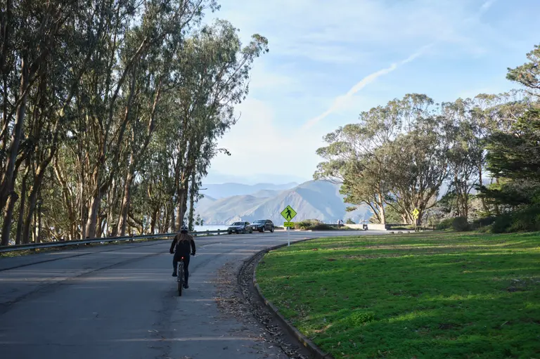 Kat biking around the apex of a curve on Washington Blvd with trees overhanging and a view towards the Marin Headlands