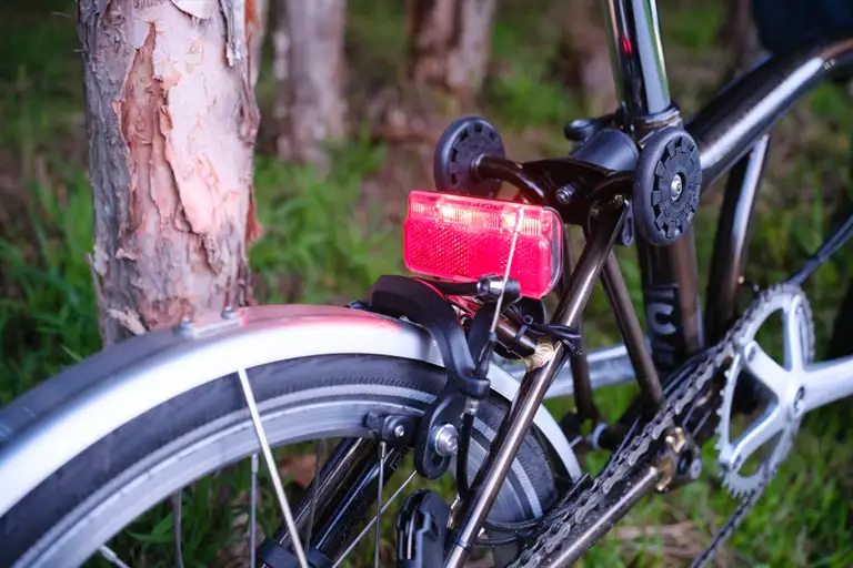 The rear of a Brompton leaned against a tree with its large rear light on.