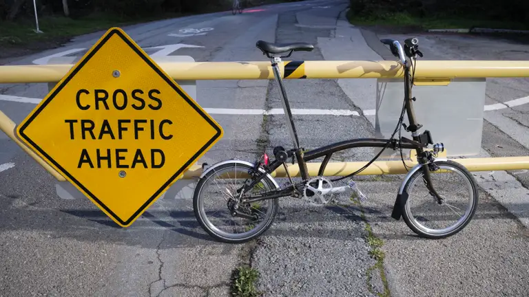 A Brompton leaning against a gate beside a traffic sign that reads 'Cross Traffic Ahead'