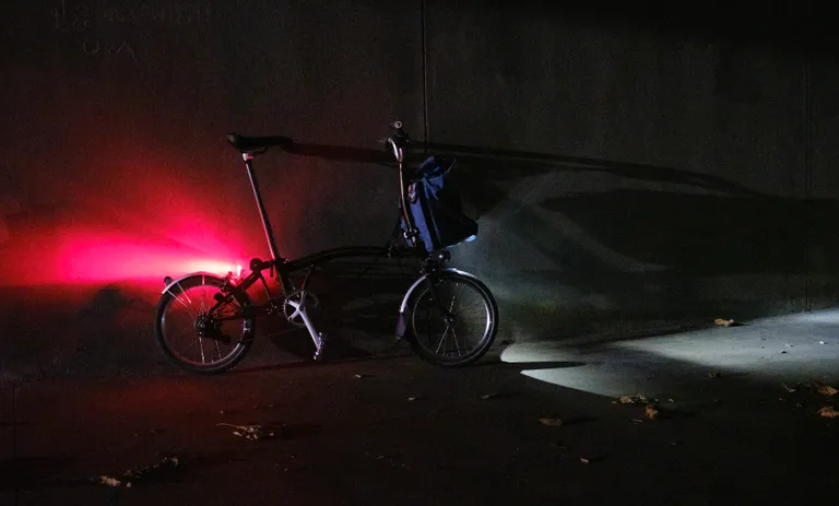 A Brompton leaned against a concrete wall in dim light with a strong rear red light and front light with tire and bag shadow.