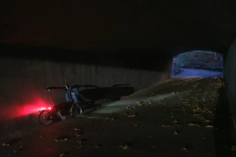 A Brompton leaning against a concrete wall in a tunnel with the opening of the tunnel in the background. 