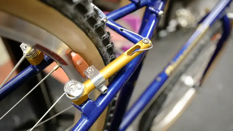 A close-up of a gold brake with the excess cable routed through its slot. It's mounted on a blue bike in a bike stand with a tanwall tire with black knobs.