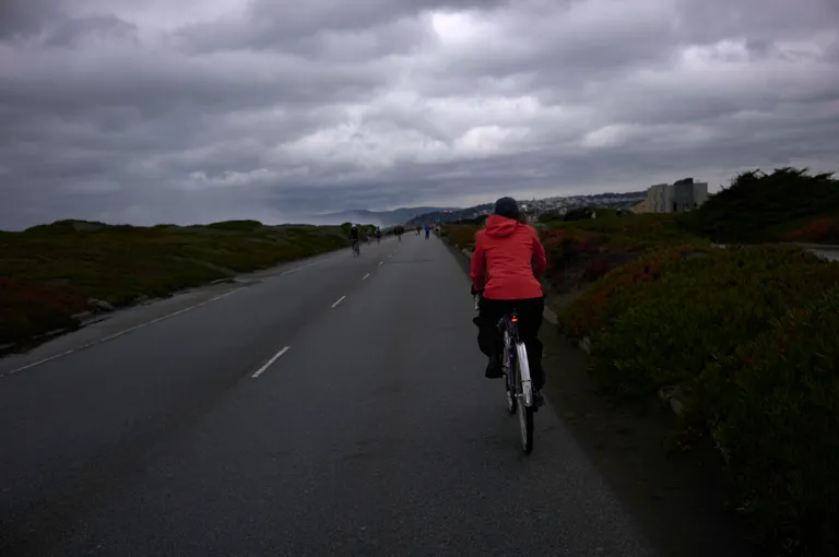 Kat in her red jacket biking down a road under very moody clouds