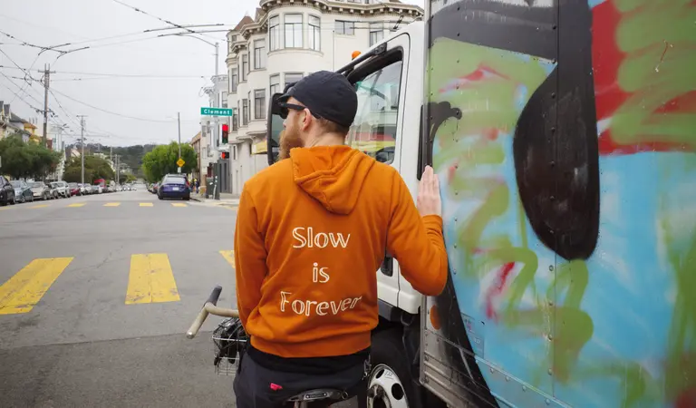 Ben on his Crust Romanceur in an orange hoodie that reads 'Slow is Forever' leaning against a colorful truck at a traffic stop.