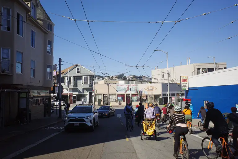 A group of cyclists waiting at the intersection of Cortland and Mission with Sutro Tower in the distance.