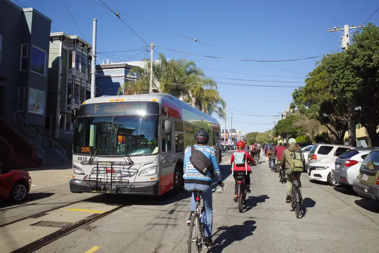 A group of cyclists on Noe St coming up to Market, riding past an F bus.