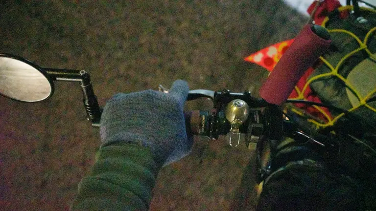 My hand in a blue wool glove on a brake lever while riding. On the left is a mirror and on the right is a bell.